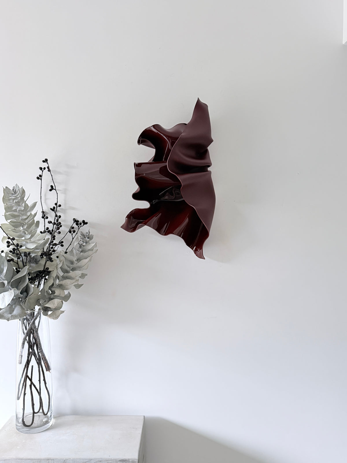 Deep Rosè, a wall sculpture by Bec Stevens in Chocolate Burgundy, on a white wall beside a vase on a plinth, showing scale