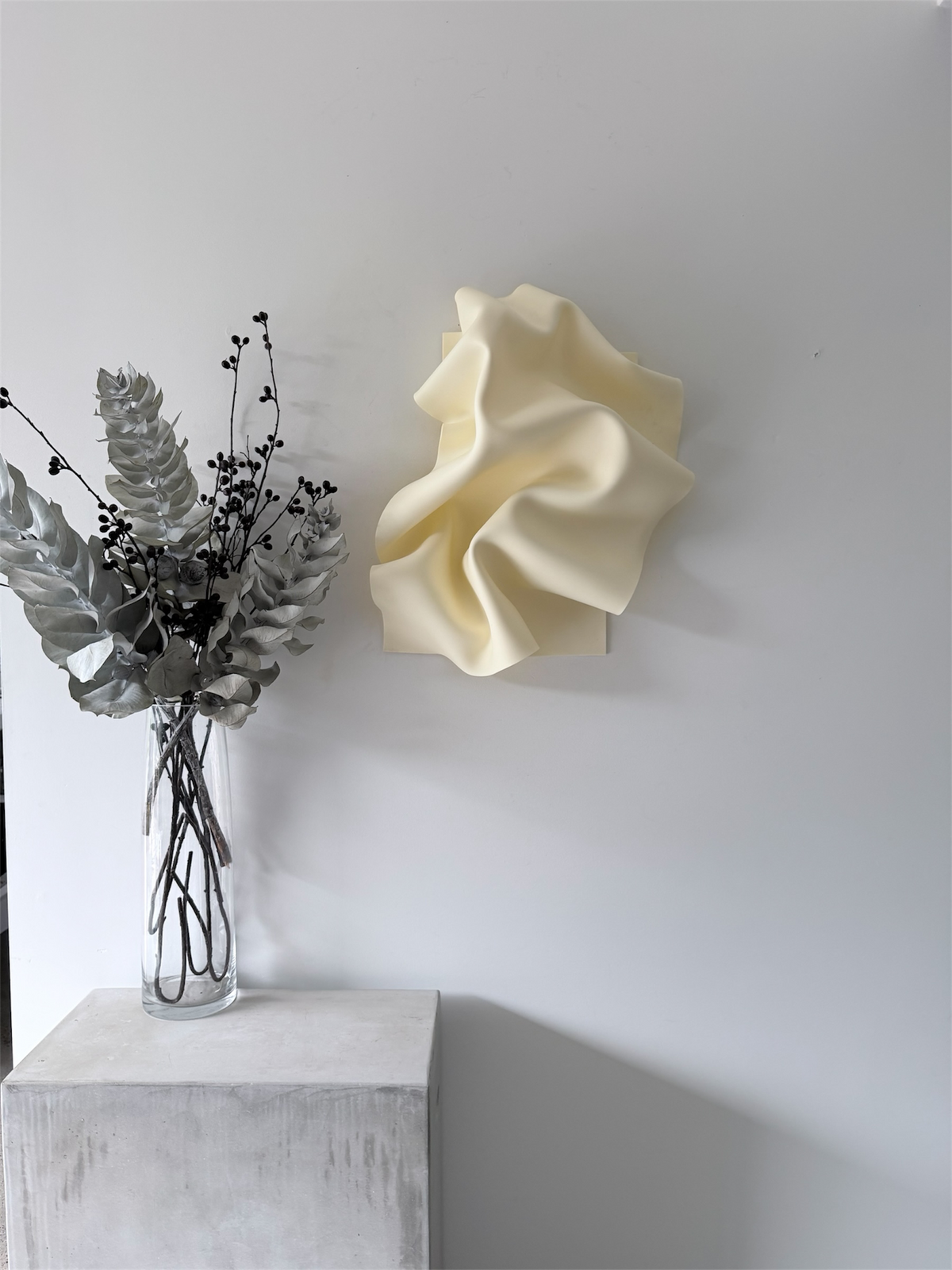 Crème de la Creme, a wall sculpture by Bec Stevens, on a white wall beside a vase on a plinth, showing scale